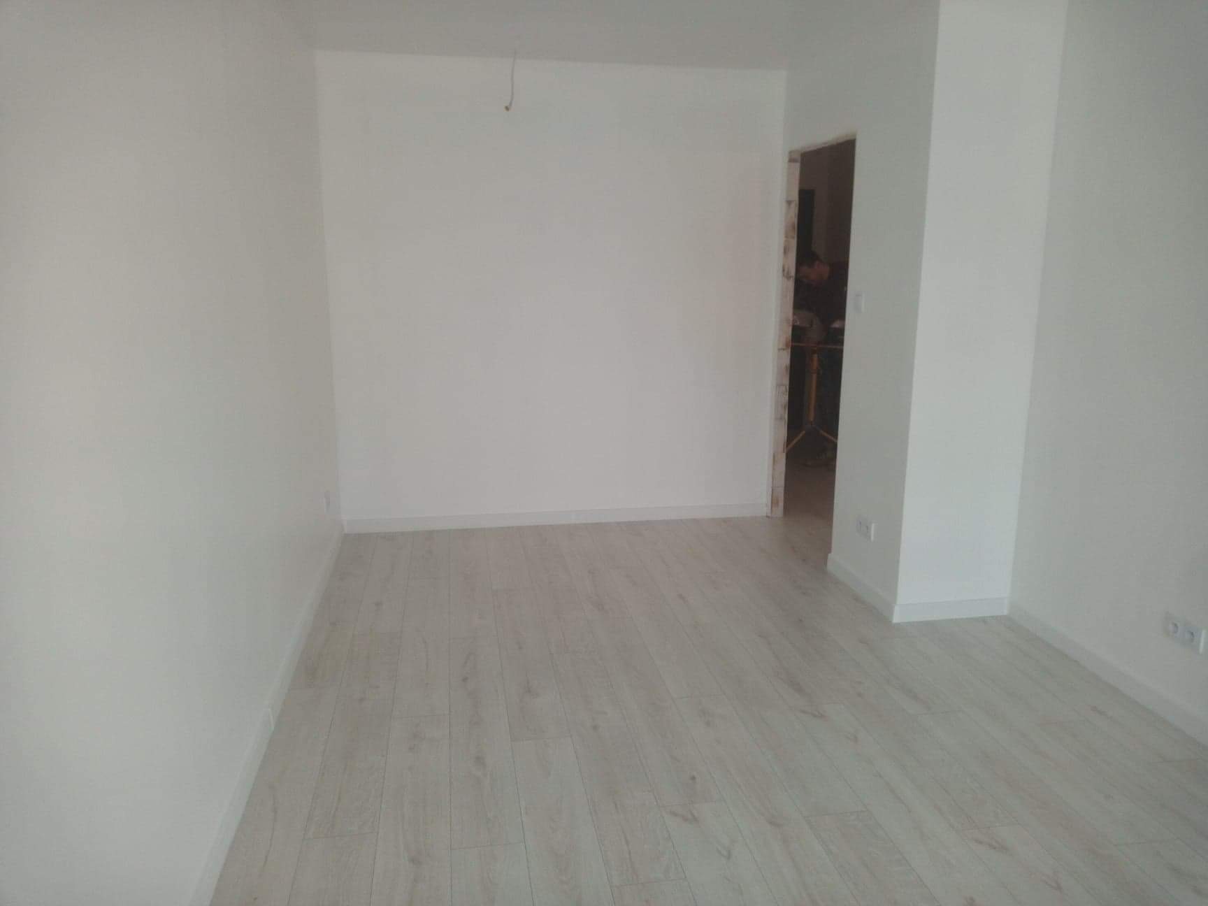 Wnętrze pokoju po malowaniu na biało, z jasnymi panelami podłogowymi i widocznym fragmentem niezabezpieczonego otworu drzwiowego w tle.