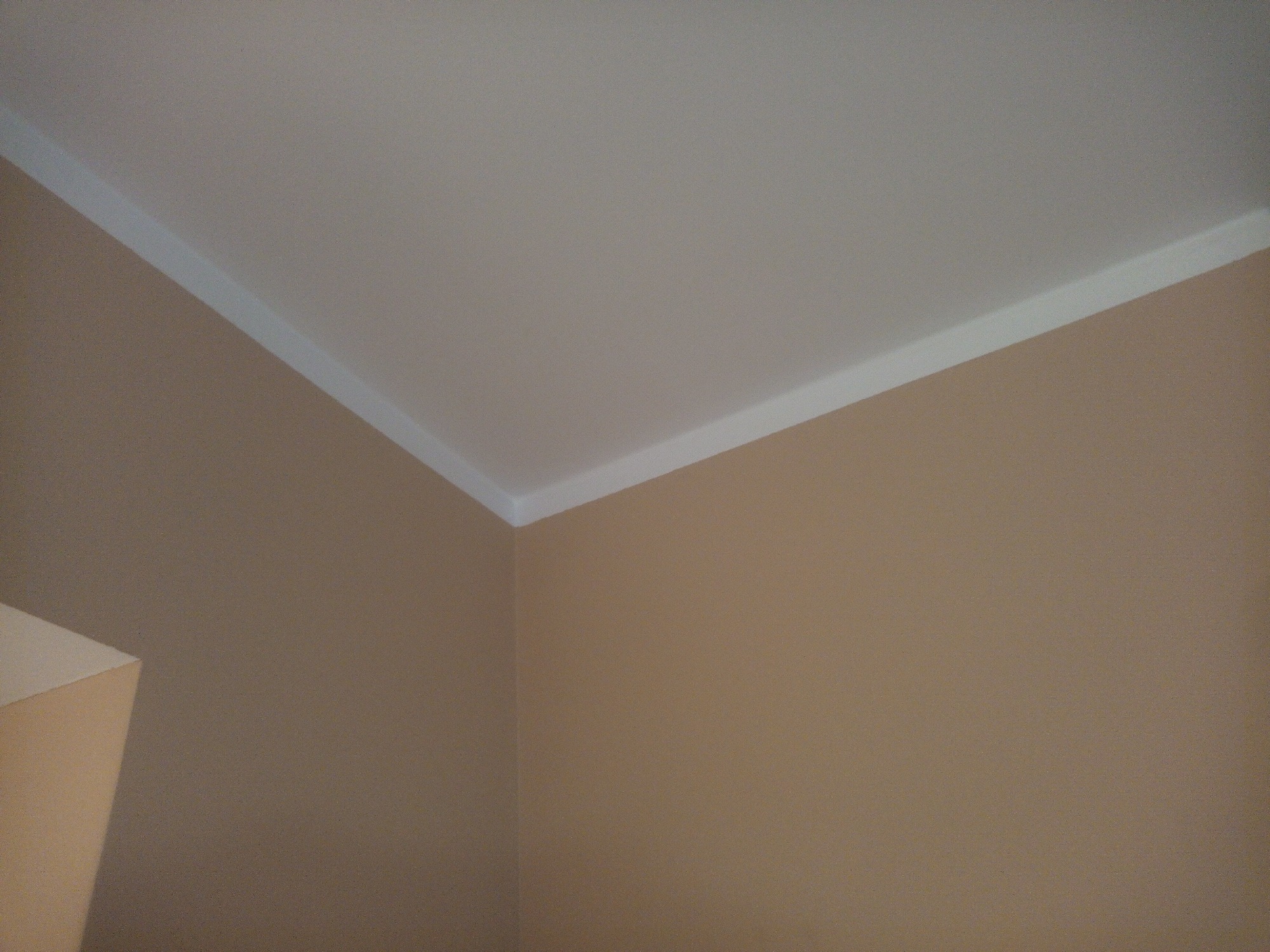 Kąt pokoju z beżowymi ścianami i białym sufitem z ozdobną listwą przysufitową.