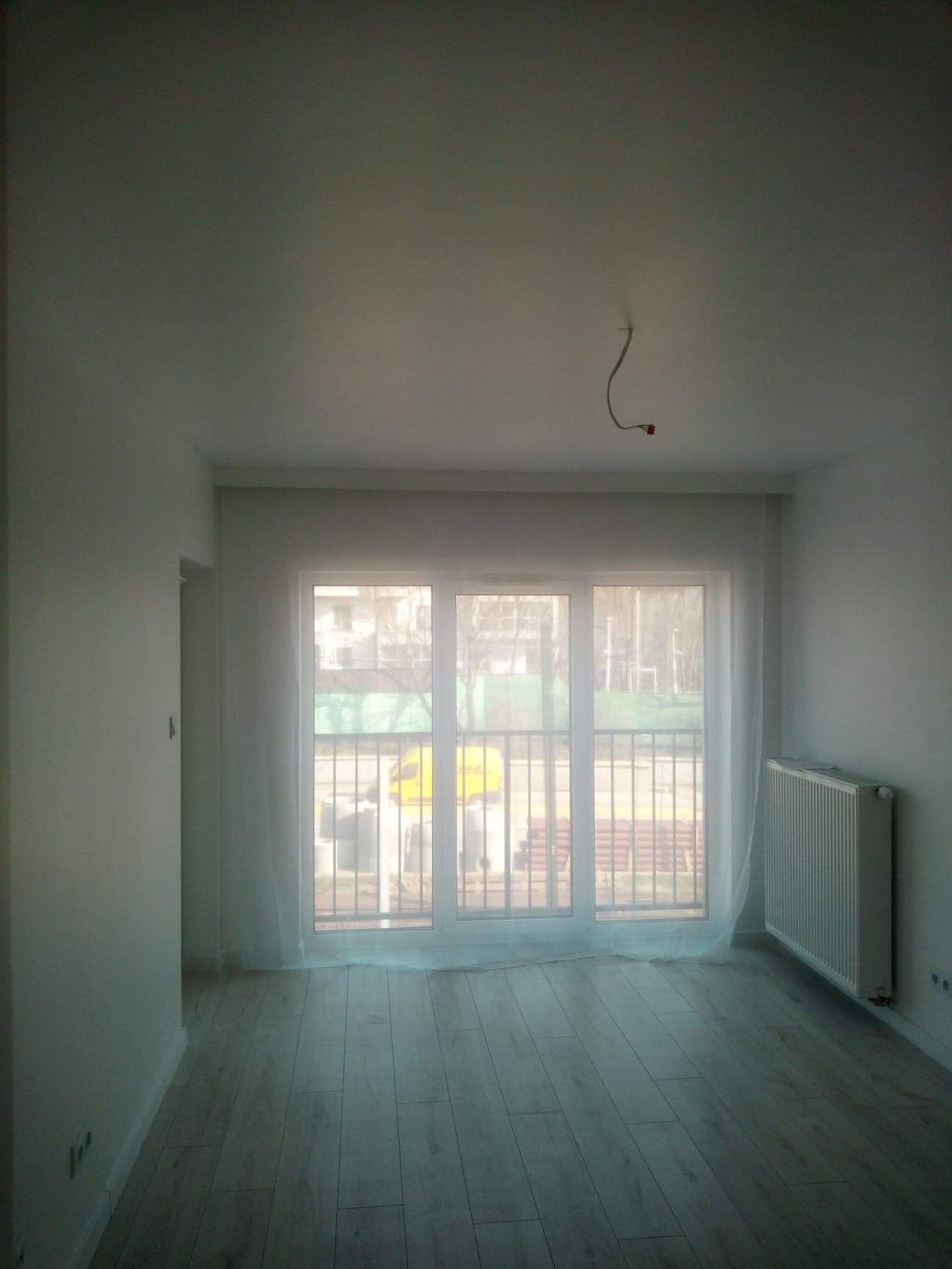 Pusty, jasny pokój po remoncie z nową podłogą z paneli, świeżo pomalowanymi ścianami i grzejnikiem pod oknem, zabezpieczonym folią. Z sufitu zwisa kabel.