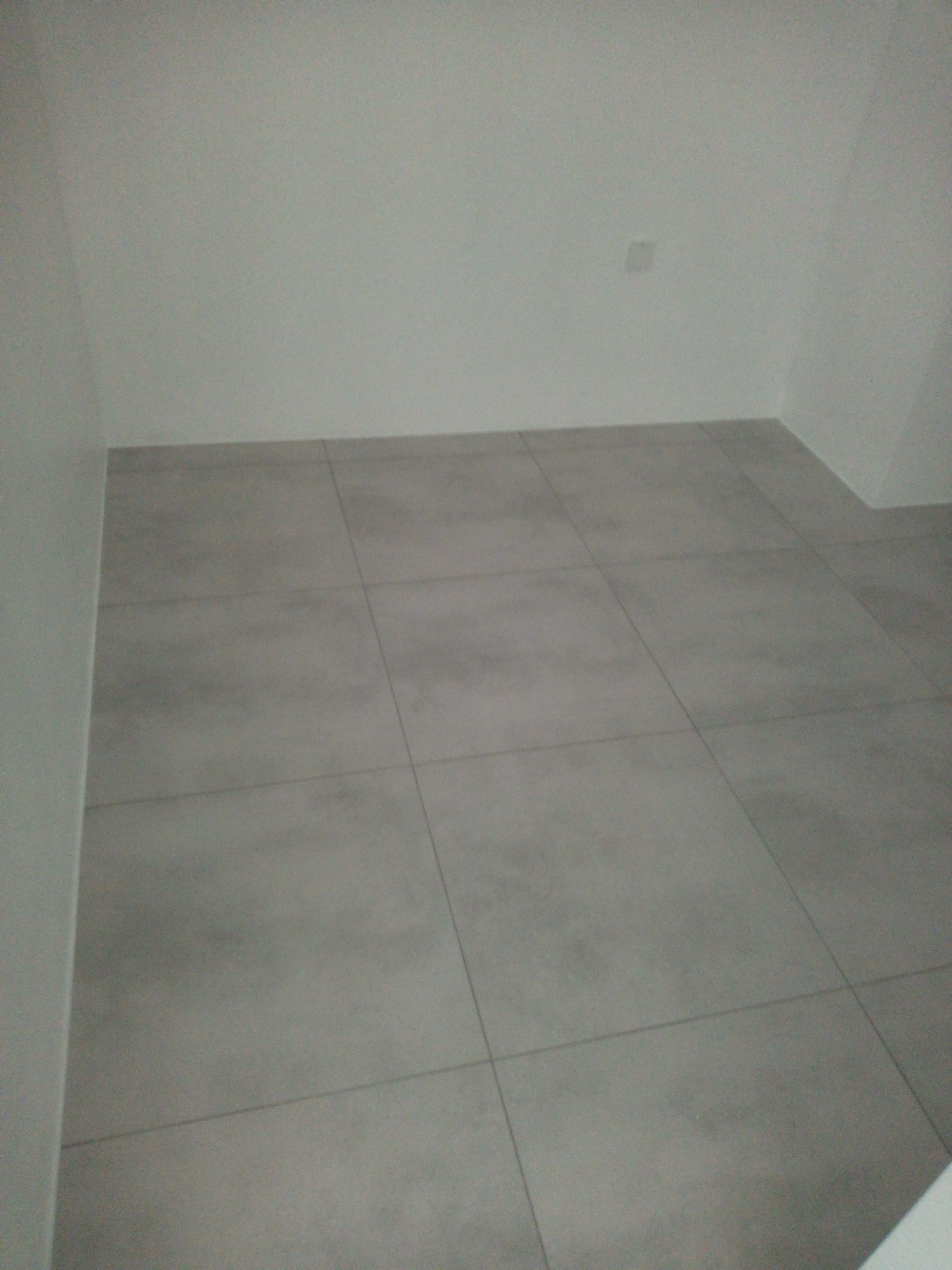 Szare płytki podłogowe ułożone w regularny wzór w rogu pokoju z białymi ścianami, widoczne spoiny między płytkami.