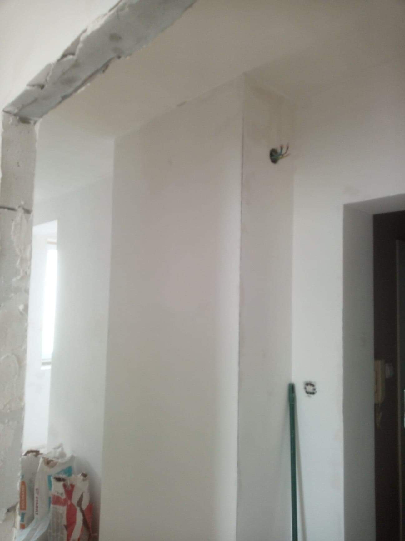 Ściana po gipsowaniu, widoczne worki z gipsem, niezakończone wykończenie framugi drzwiowej i wystające kable elektryczne.