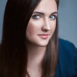 Portret kobiety z długimi brązowymi włosami i niebieskimi oczami, spoglądającej w górę, na jednolitym szarym tle, ubrana w ciemnoniebieską bluzkę.