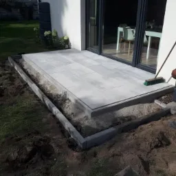 Montaż tarasu z szarych płyt kamiennych, widoczne obrzeża i przygotowane podłoże, pracownik zamiata powierzchnię. Widok na okno tarasowe i ogród.