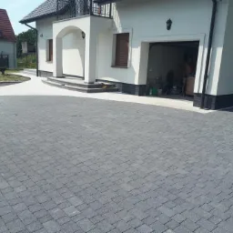 Ułożona szara kostka brukowa prowadząca do białego domu z garażem i podjazdem z jasnych płyt, widoczny fragment trawnika z placem zabaw.