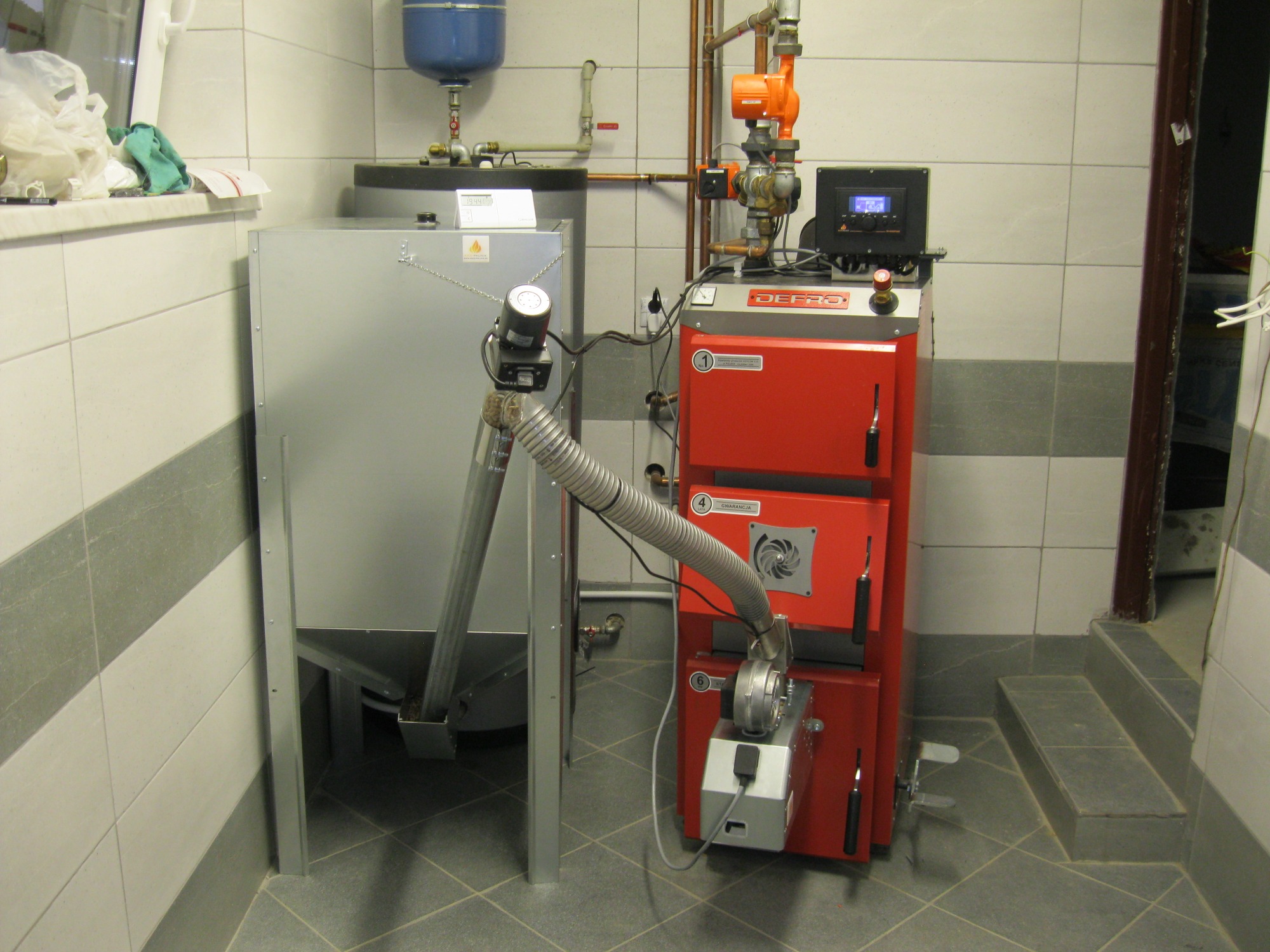 Czerwony piec z podajnikiem na ekogroszek marki DEFRO, połączony elastycznym przewodem z zasobnikiem na paliwo, zainstalowany w kotłowni wyłożonej płytkami ceramicznymi.
