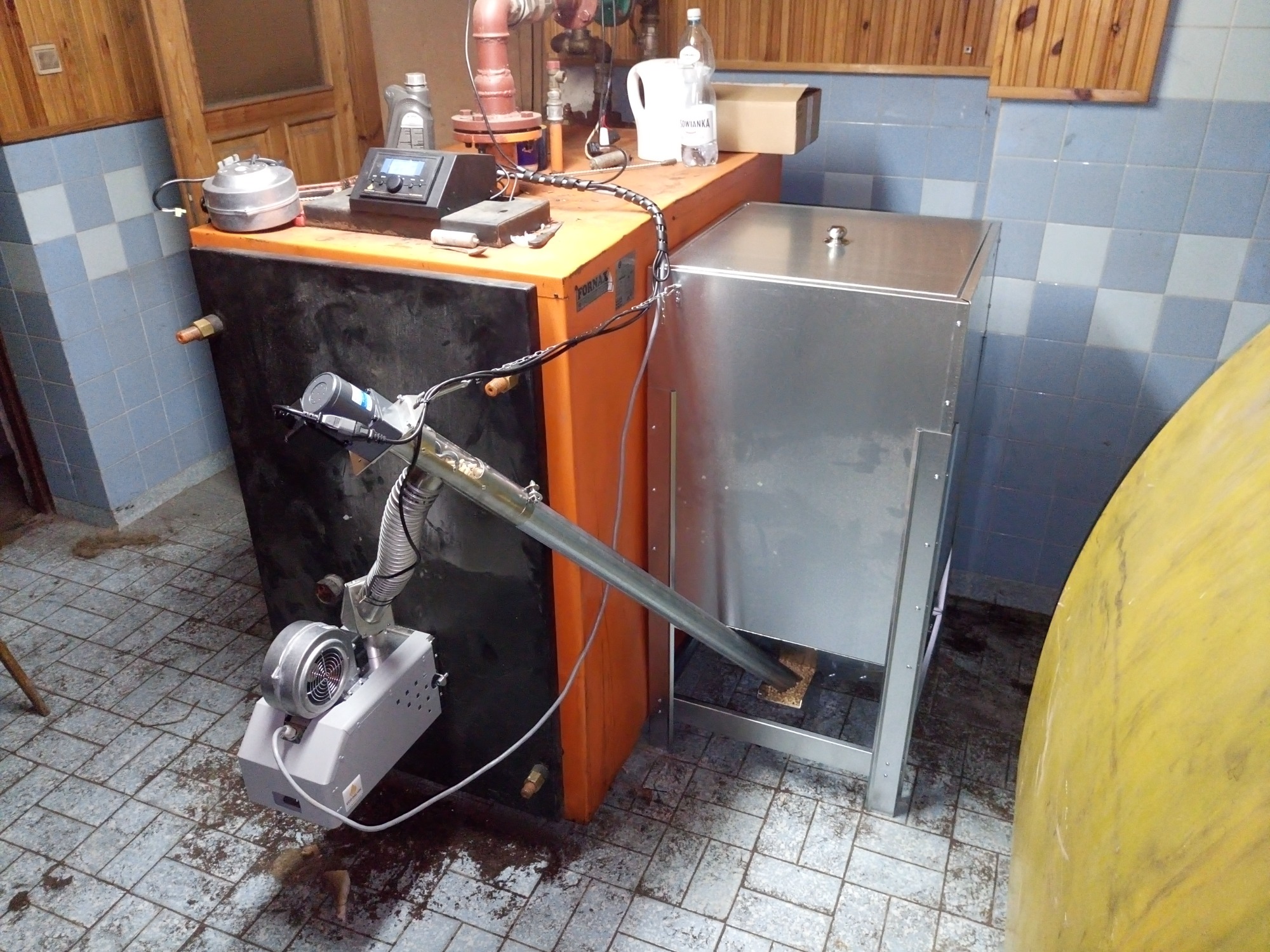 Pomarańczowo-czarny piec z podajnikiem ślimakowym i metalową obudową, widoczne elementy instalacji, otoczenie w płytkach, lekki bałagan na podłodze.