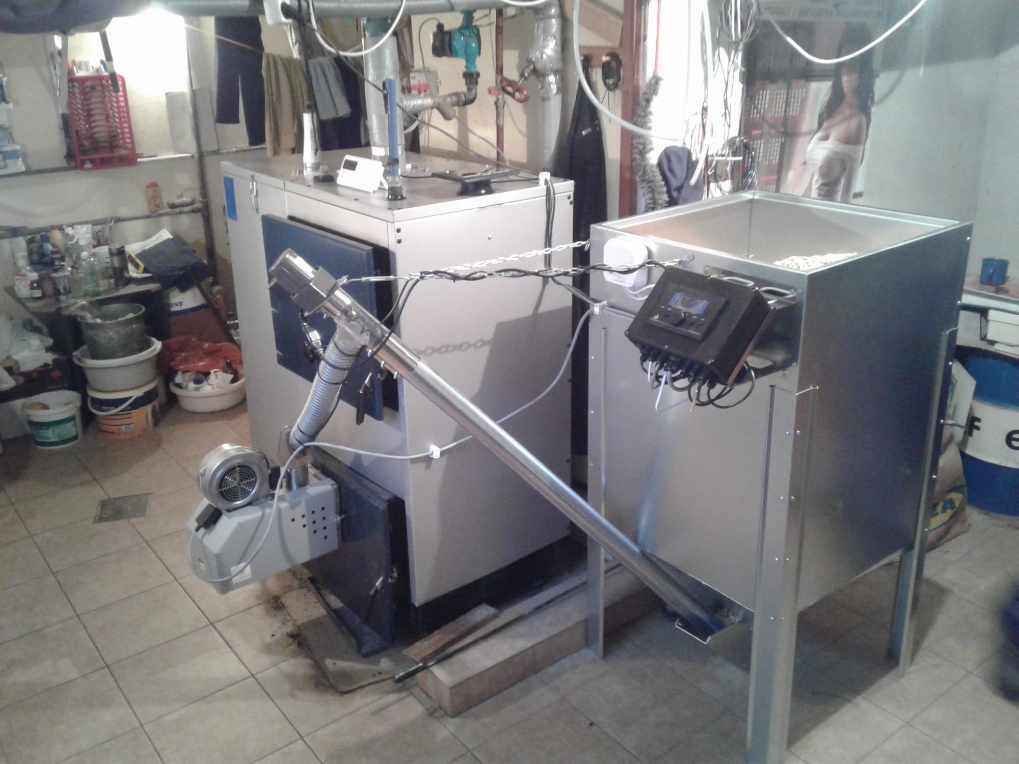 Automatyczny podajnik peletu połączony z kotłem w kotłowni o standardowym wykończeniu, widoczne rury i osprzęt instalacji.