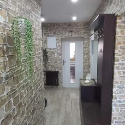 Wąski korytarz z okładziną ścienną imitującą kamień, jasnymi panelami podłogowymi i ciemnobrązową szafką z lustrem, wisząca roślina w doniczce.