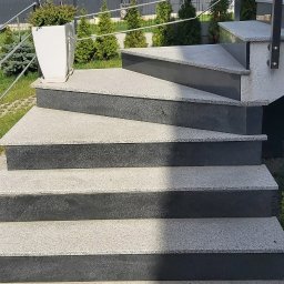 Bud-Mar Marcin Kołtonowski - Zewnętrzne schody z szarego kamienia z metalową balustradą, ozdobione białą donicą z zielonymi roślinami, na tle zieleni i zabudowań mieszkalnych.