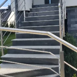 Zewnętrzne schody wejściowe z szarego granitu z kontrastującymi, ciemniejszymi podstopnicami i nowoczesną balustradą ze stali nierdzewnej, prowadzące do ciemnoszarych drzwi wejściowych.