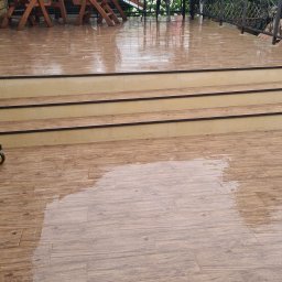 Bud-Mar Marcin Kołtonowski - Wypłytkowany, mokry taras z drewnianymi meblami ogrodowymi i schodami; widoczne odbicia wody na powierzchni płytek imitujących drewno.