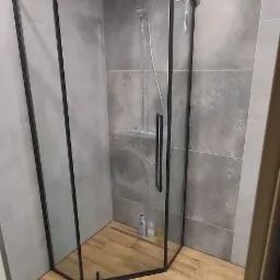 Kabina prysznicowa z czarną ramą w wyremontowanej łazience, drewnopodobne płytki na podłodze, szare płytki na ścianach z delikatnym wzorem, widoczne dwa punkty oświetleniowe na suficie.