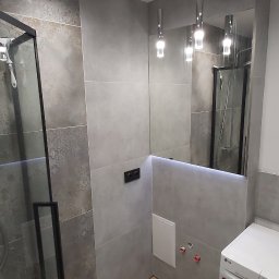 Bud-Mar Marcin Kołtonowski - Nowoczesna łazienka z szarymi płytkami o zróżnicowanej fakturze, kabiną prysznicową z czarnymi ramami, lustrem z podświetleniem LED, drewnopodobną podłogą i pralką LG w rogu.