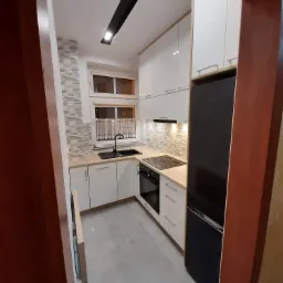 Wnętrze małej, nowocześnie urządzonej kuchni z białymi szafkami, czarnym zlewem i lodówką, oraz płytkami imitującymi kamień na ścianie nad blatem, widziane zza framugi drzwi.