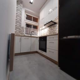 Bud-Mar Marcin Kołtonowski - Nowoczesna kuchnia z białymi szafkami, czarnym piekarnikiem i lodówką, szarymi płytkami na ścianie nad blatem i podświetleniem LED pod szafkami wiszącymi.