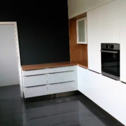 Nowoczesna kuchnia z białymi szafkami, drewnianym blatem i czarną ścianą akcentującą, widok na błyszczące, ciemne płytki podłogowe.