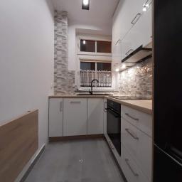 Bud-Mar Marcin Kołtonowski - Nowoczesna, wąska kuchnia z białymi, błyszczącymi szafkami, drewnianym blatem, szarą podłogą i dekoracyjną mozaiką na ścianie nad blatem, oświetlona czarną lampą sufitową.