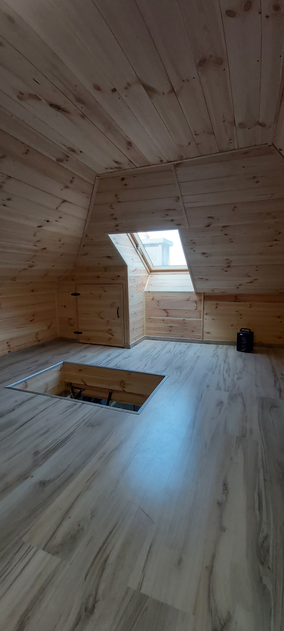 Drewniane wykończenie poddasza z oknem dachowym, widoczne panele podłogowe i otwór w podłodze z mechanizmem podnoszenia.