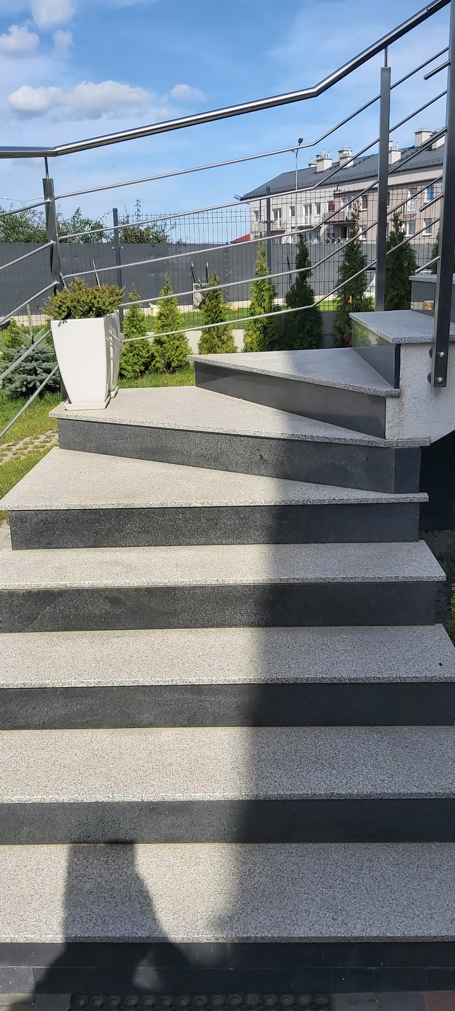 Zewnętrzne schody z szarego kamienia z metalową balustradą, ozdobione białą donicą z zielonymi roślinami, na tle zieleni i zabudowań mieszkalnych.