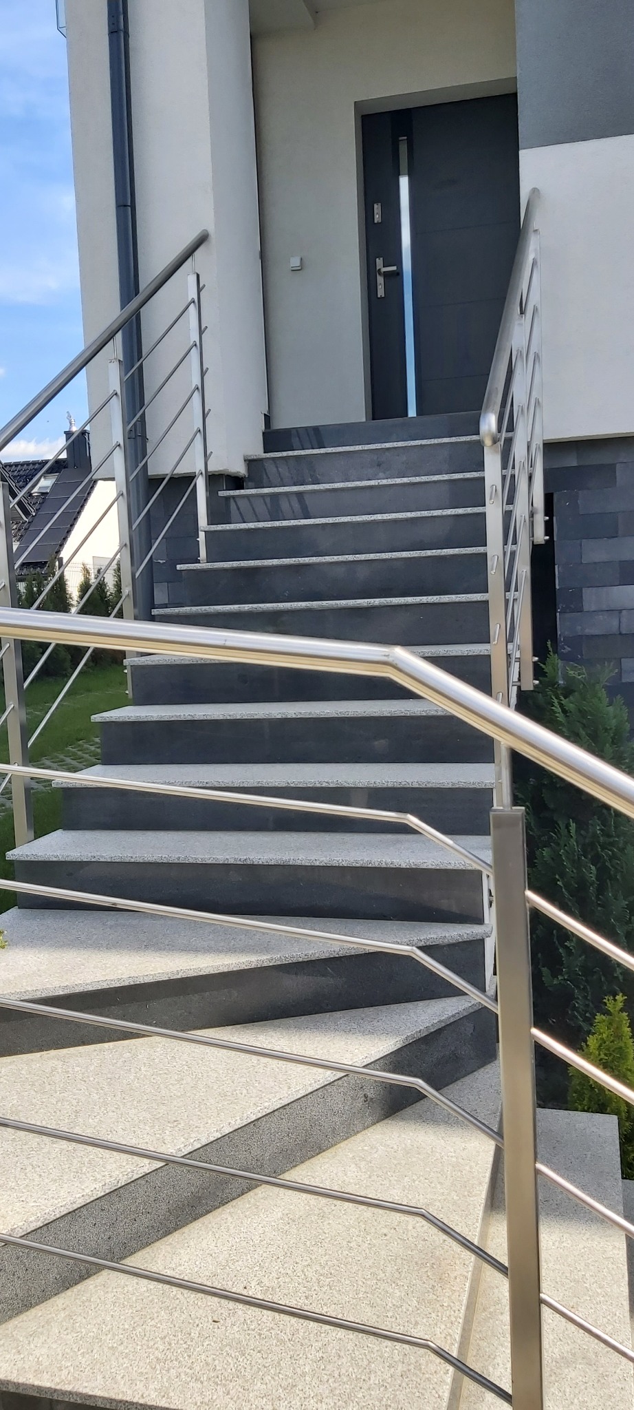 Zewnętrzne schody wejściowe z szarego granitu z kontrastującymi, ciemniejszymi podstopnicami i nowoczesną balustradą ze stali nierdzewnej, prowadzące do ciemnoszarych drzwi wejściowych.