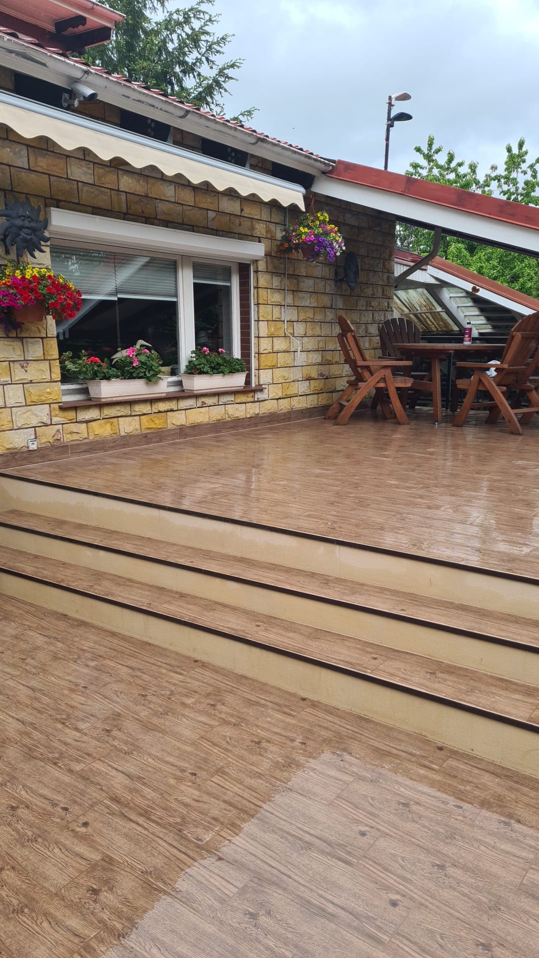 Zewnętrzny taras z drewnopodobnych płytek ceramicznych, trzy stopnie prowadzące do przestrzeni z drewnianym stołem i krzesłami, widok na okno z kwiatami w donicach, murowana ściana z markizą.