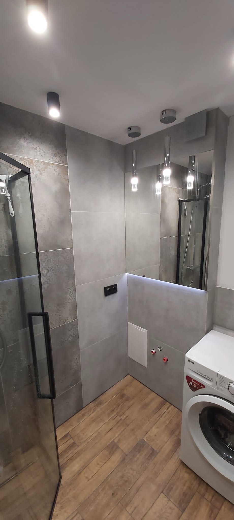 Nowoczesna łazienka w odcieniach szarości z drewnopodobną podłogą, kabina prysznicowa z czarnymi profilami, lustro z podświetleniem LED i pralka LG.