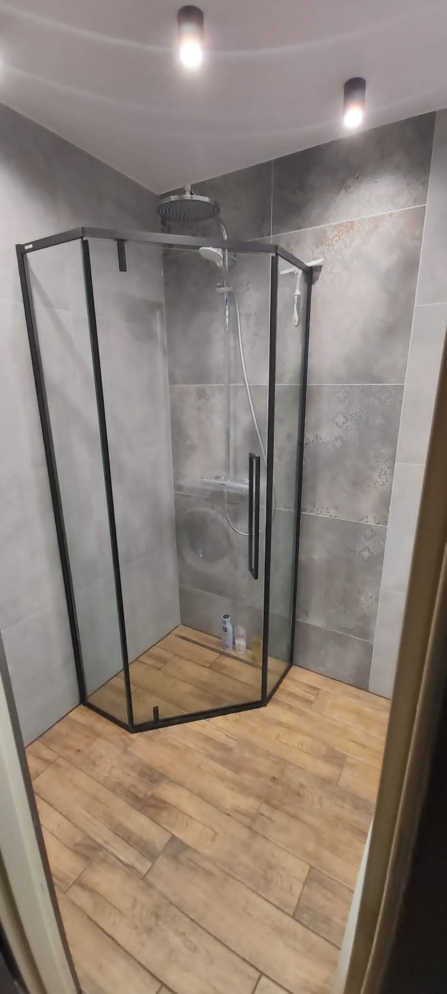 Kabina prysznicowa z czarną ramą w wyremontowanej łazience, drewnopodobne płytki na podłodze, szare płytki na ścianach z delikatnym wzorem, widoczne dwa punkty oświetleniowe na suficie.