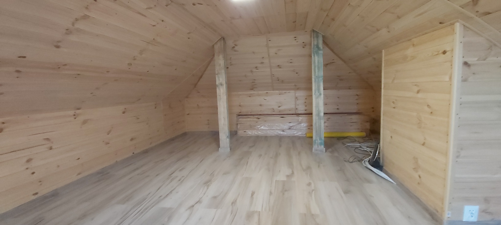 Wykończone poddasze z drewnianą boazerią na ścianach i suficie, jasne panele podłogowe, widoczne słupy konstrukcyjne i materiały budowlane w folii.