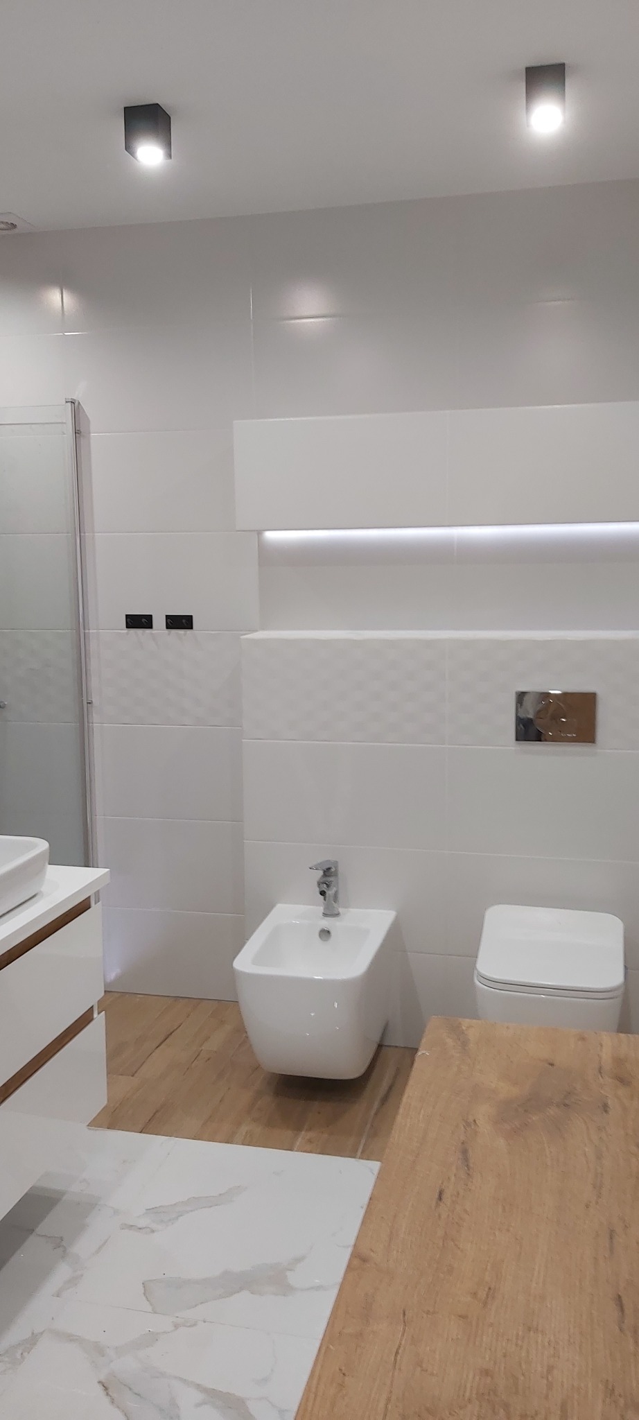 Nowoczesna łazienka z białymi płytkami o różnych fakturach, podświetlaną wnęką, wiszącym bidetem i toaletą oraz drewnopodobną podłogą.