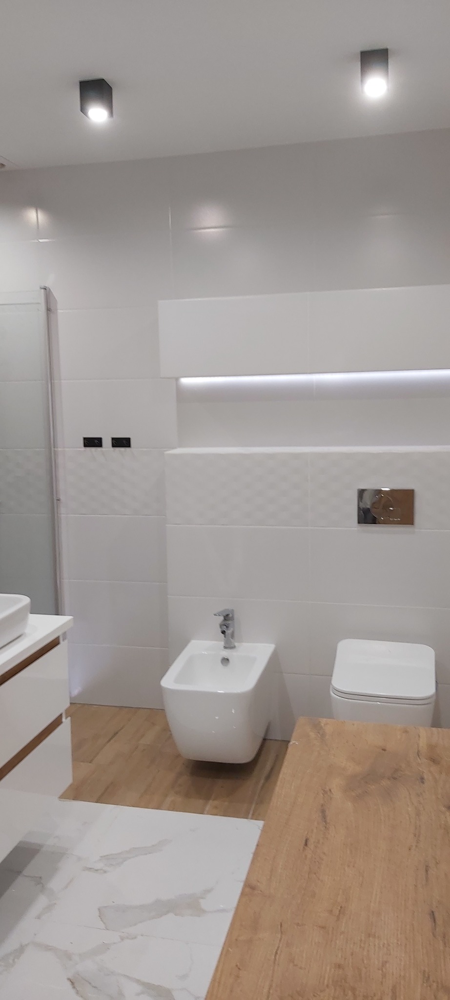 Nowoczesna łazienka z białymi płytkami o różnej fakturze, wiszącą miską WC, bidetem i drewnopodobną podłogą, oświetlona dwoma punktowymi lampami sufitowymi.