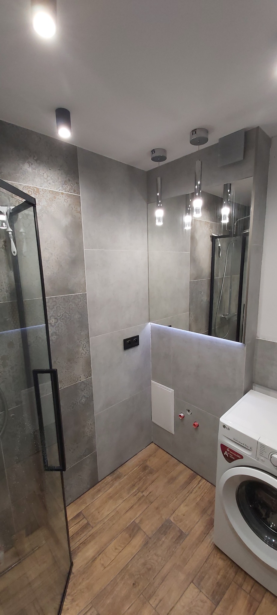 Nowoczesna łazienka z szarymi płytkami o zróżnicowanej fakturze, kabiną prysznicową z czarnymi ramami, lustrem z podświetleniem LED, drewnopodobną podłogą i pralką LG w rogu.