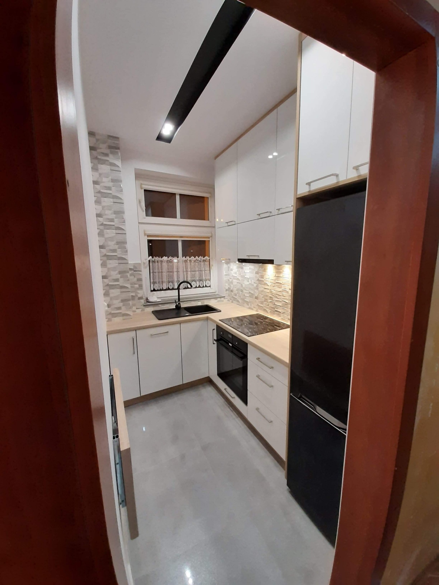 Wnętrze małej, nowocześnie urządzonej kuchni z białymi szafkami, czarnym zlewem i lodówką, oraz płytkami imitującymi kamień na ścianie nad blatem, widziane zza framugi drzwi.