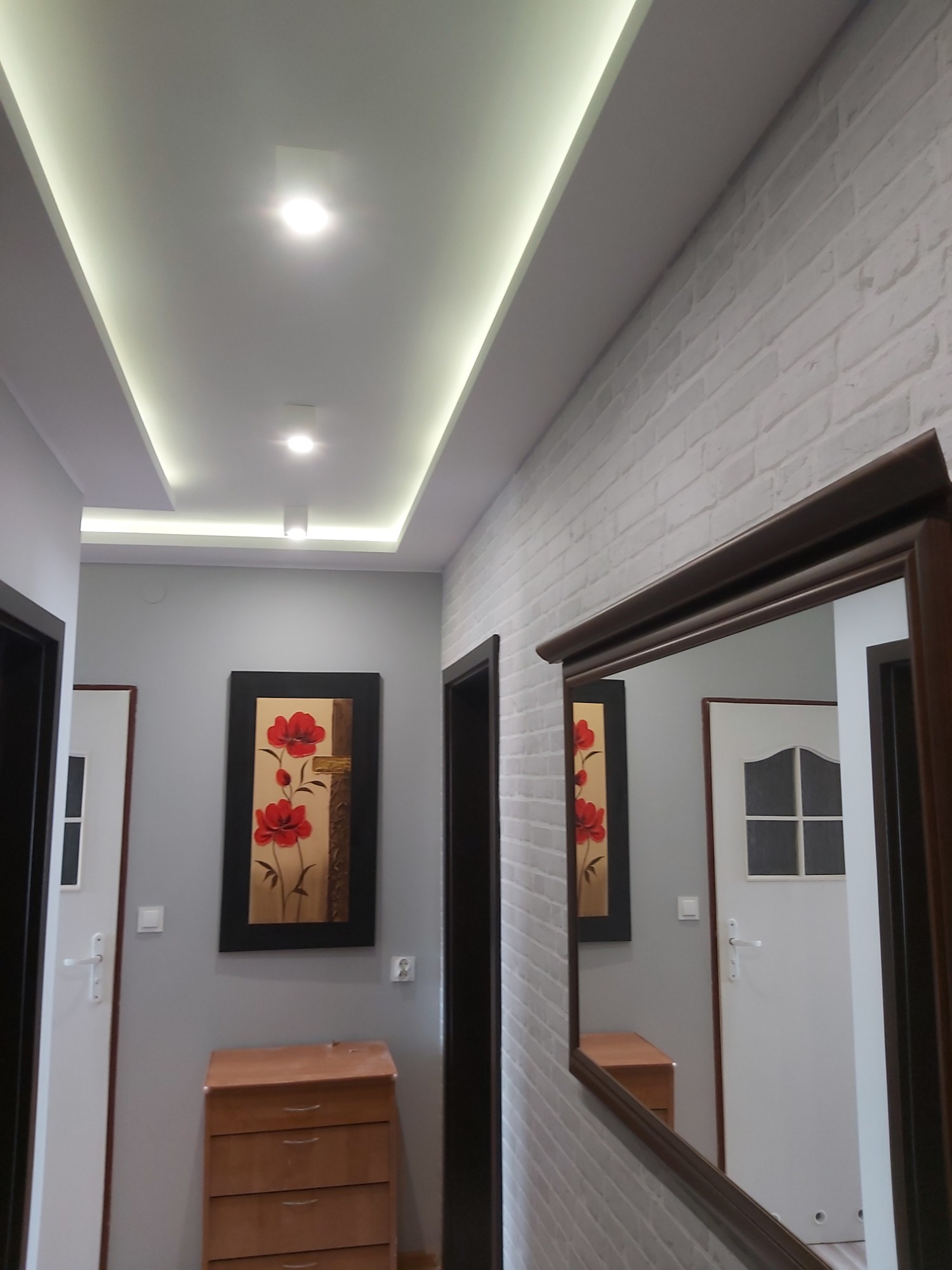 Korytarz po remoncie z oświetleniem LED w suficie, imitacją cegły na ścianie, lustrem w brązowej ramie i obrazem z czerwonymi kwiatami.