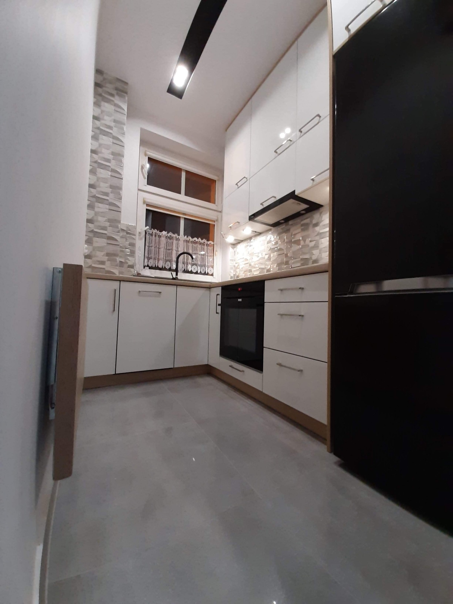 Nowoczesna kuchnia z białymi szafkami, czarnym piekarnikiem i lodówką, szarymi płytkami na ścianie nad blatem i podświetleniem LED pod szafkami wiszącymi.