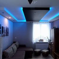 Salon z narożną sofą i dekoracyjnym sufitem podwieszanym z niebieskim oświetleniem LED, ściana z cegieł, obrazy na ścianie, widok na okno.