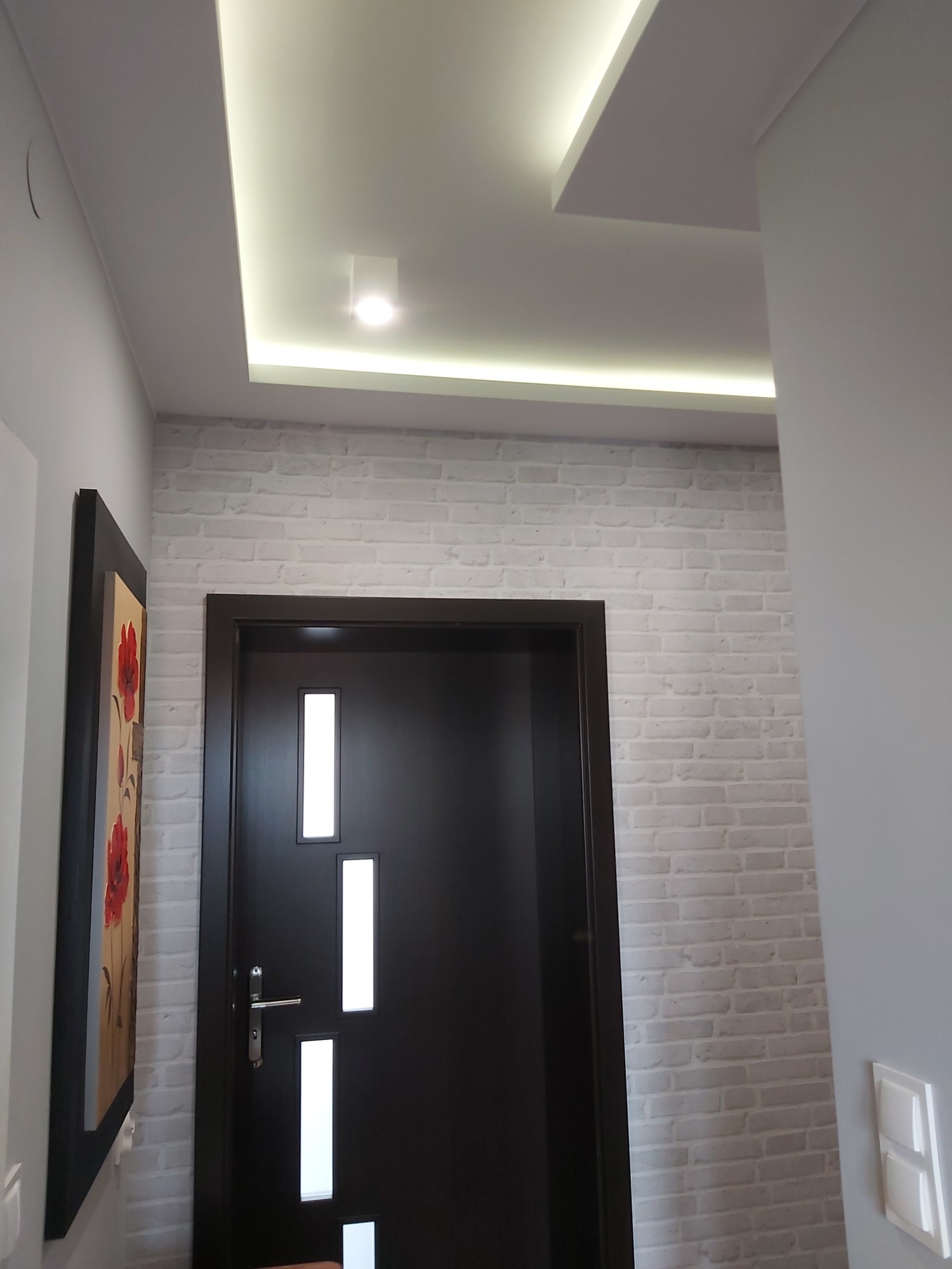 Wnętrze: czarne drzwi z trzema pionowymi przeszkleniami na tle ściany z białej cegły, oświetlenie sufitowe LED w suficie podwieszanym, obraz z motywem kwiatowym po lewej stronie.