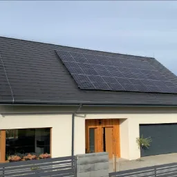 Nowoczesny dom jednorodzinny z panelami słonecznymi na dachu pokrytym grafitową dachówką, jasna elewacja, drewniane drzwi wejściowe i brama garażowa w kolorze antracytowym.