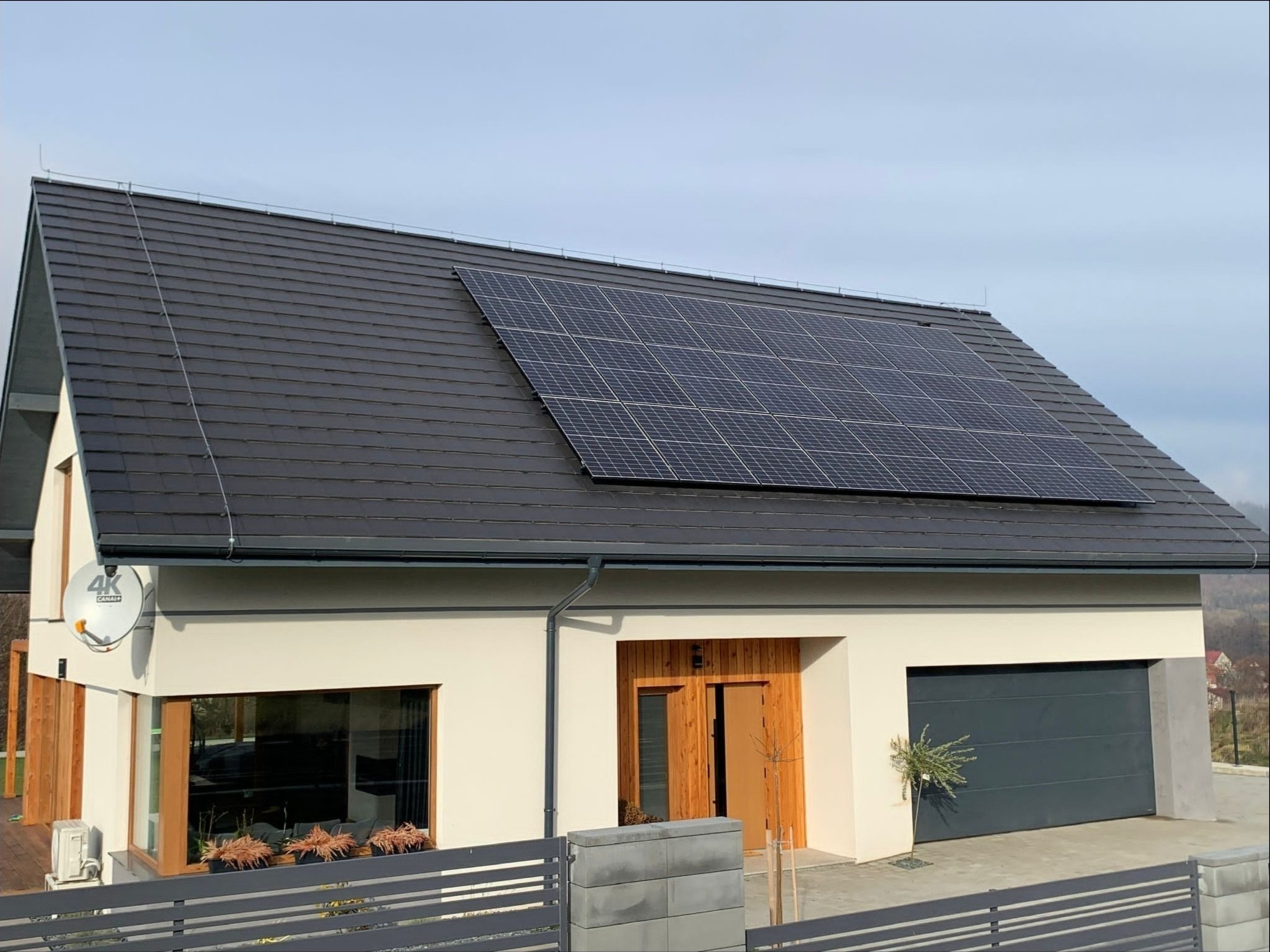 Nowoczesny dom jednorodzinny z panelami słonecznymi na dachu pokrytym grafitową dachówką, jasna elewacja, drewniane drzwi wejściowe i brama garażowa w kolorze antracytowym.