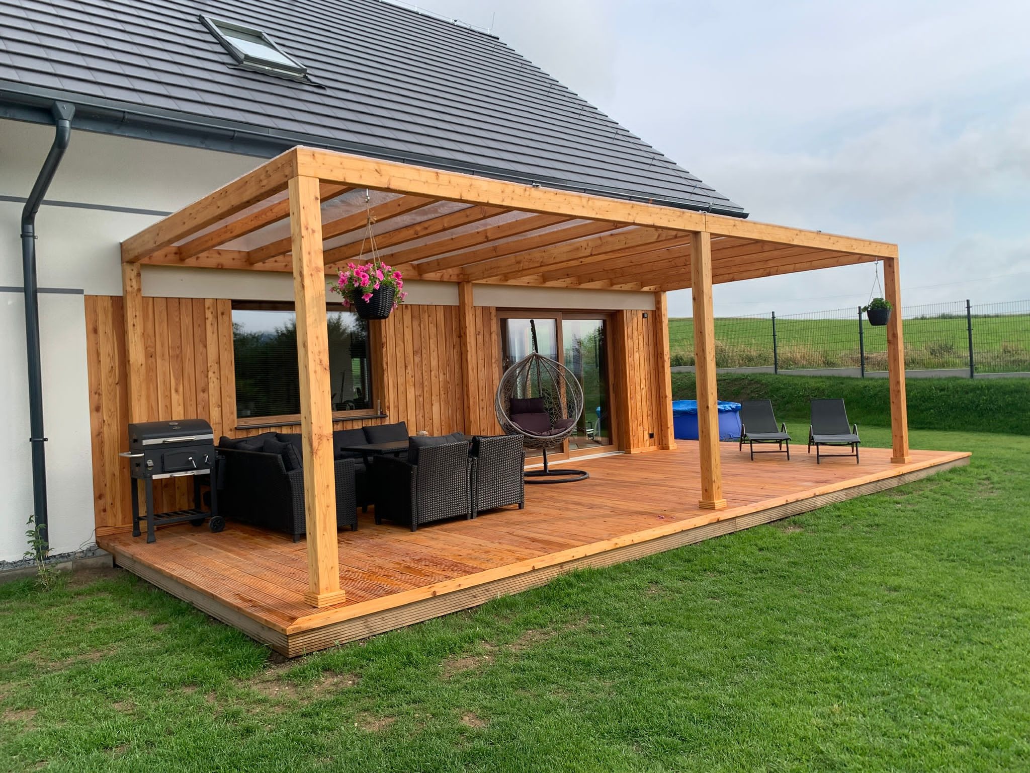 Drewniana konstrukcja zadaszenia tarasu przy domu z meblami ogrodowymi i grillem, wiszące doniczki z kwiatami, widok na trawnik i ogrodzenie.