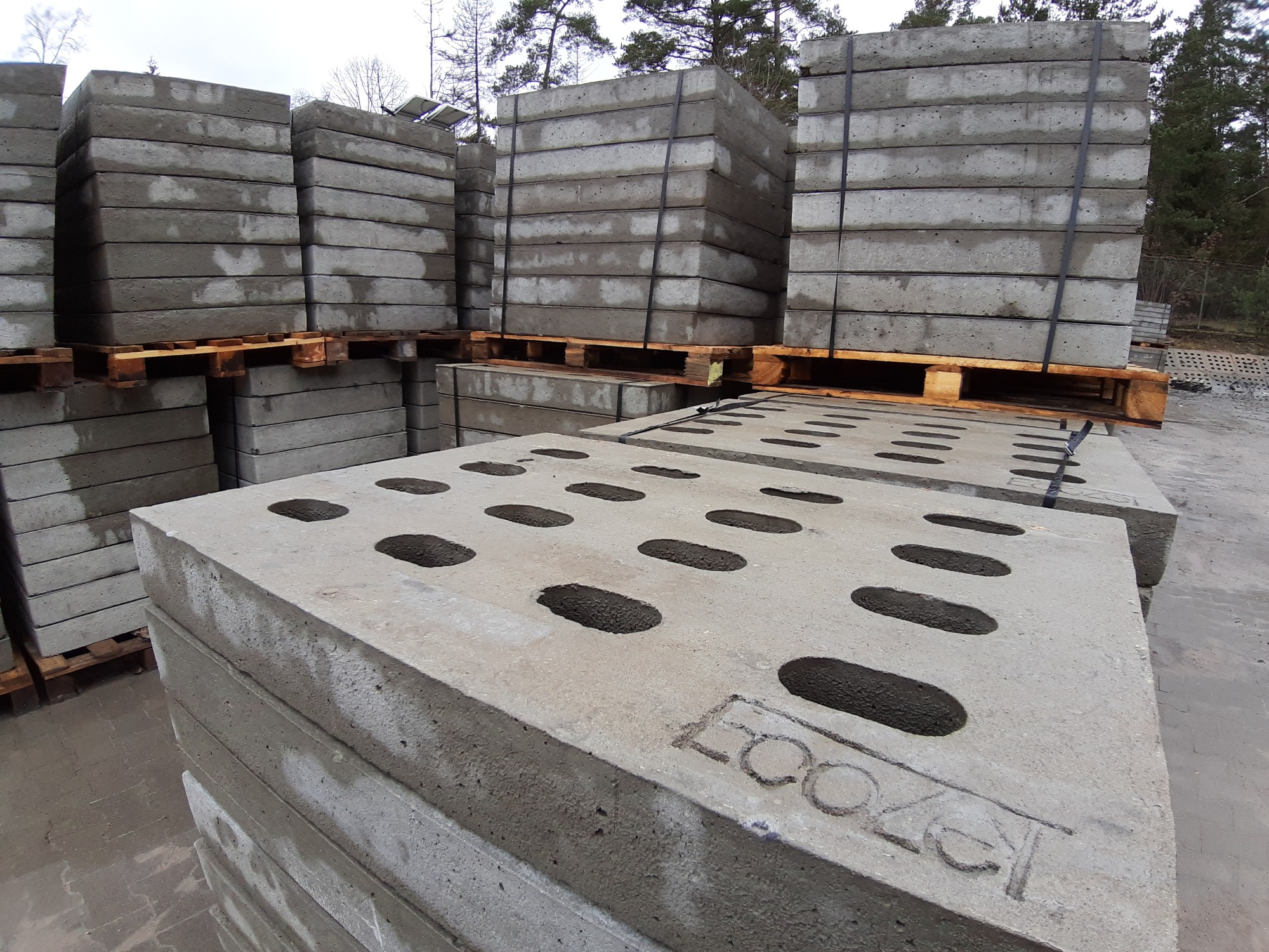 Stosy betonowych płyt na drewnianych paletach, z widocznymi otworami i napisem 'LOOZER' na jednej z płyt, tło stanowią drzewa.