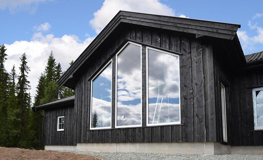 Ciemny, drewniany dom szkieletowy z dużym trójdzielnym oknem, w którym odbija się błękitne niebo z chmurami; widoczna drabina oparta o ścianę.