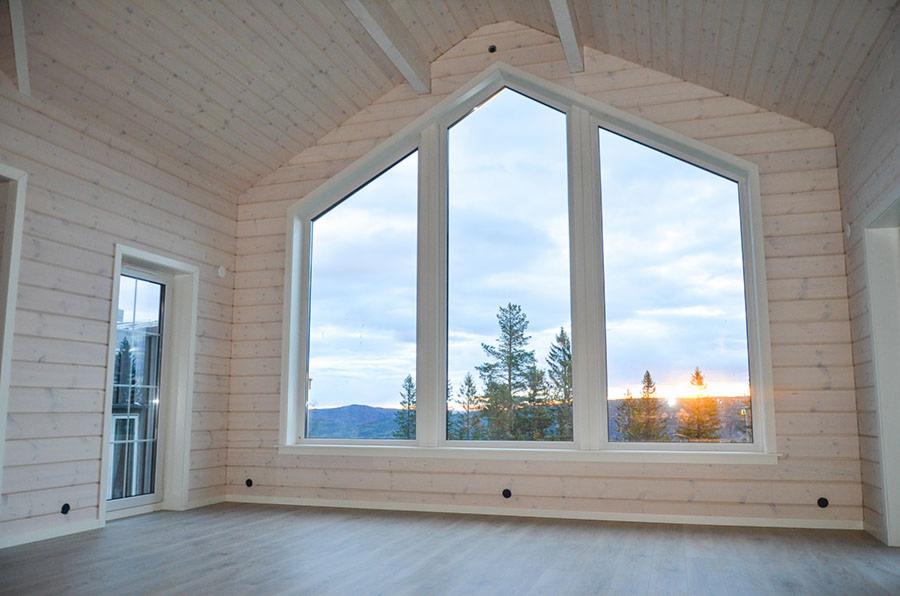 Jasne wnętrze z drewnianą boazerią, trójkątne okno z widokiem na zachód słońca nad lasem i wzgórzami, szare panele podłogowe, widoczne punkty elektryczne w ścianie.