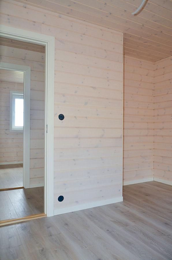 Wnętrze z wykończonymi ścianami z jasnego drewna, widoczne gniazdko elektryczne i otwarte drzwi do kolejnego pomieszczenia, podłoga laminowana.