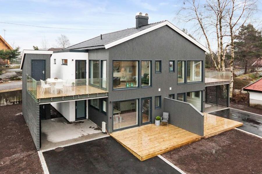 Nowoczesny dom szkieletowy z szarą elewacją, drewnianym tarasem i balkonem z przeszkloną balustradą, widok z zewnątrz w pochmurny dzień.