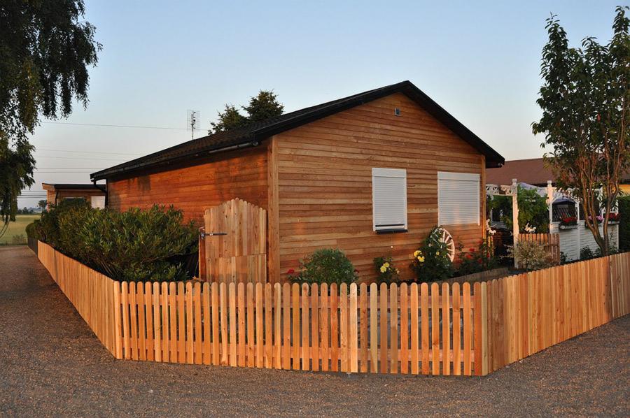 Całoroczny dom drewniany z jasną elewacją, ogrodzony niskim płotem z drewnianych sztachet, widok z zewnątrz na ogród.