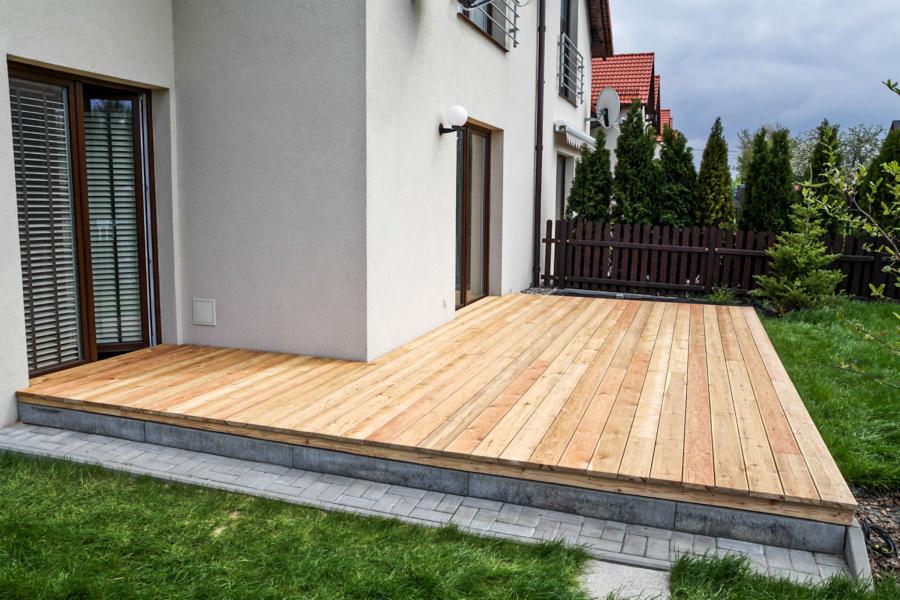 Nowoczesny taras z jasnego drewna przy białym domu, widoczne przesuwne drzwi tarasowe, szara podmurówka i kostka brukowa, zadbany trawnik.