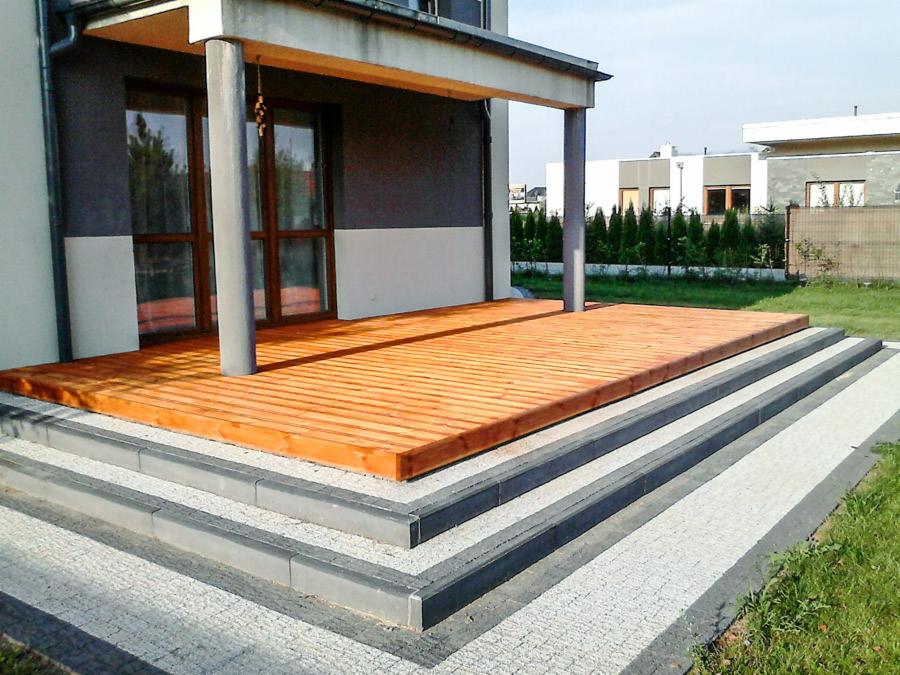 Drewniany taras z desek w kolorze miodowym, przylegający do szarego domu z kolumnami, z widocznymi schodami z kostki brukowej i zielonym trawnikiem w tle.