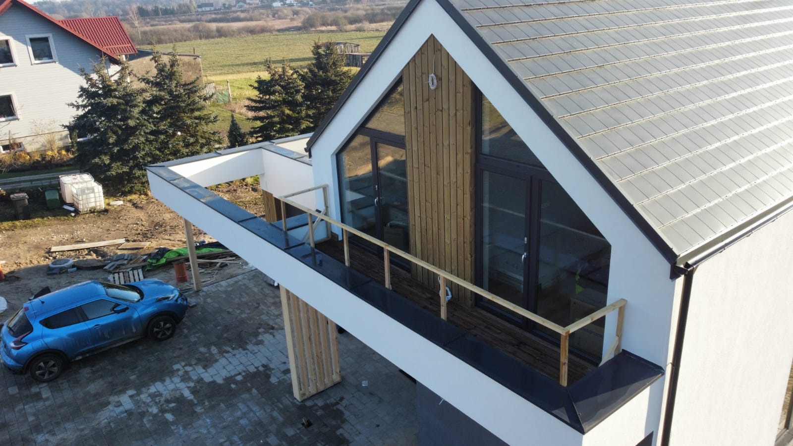 Nowoczesny dom z drewnianą elewacją, widok z góry, z balkonem i zaparkowanym niebieskim samochodem.