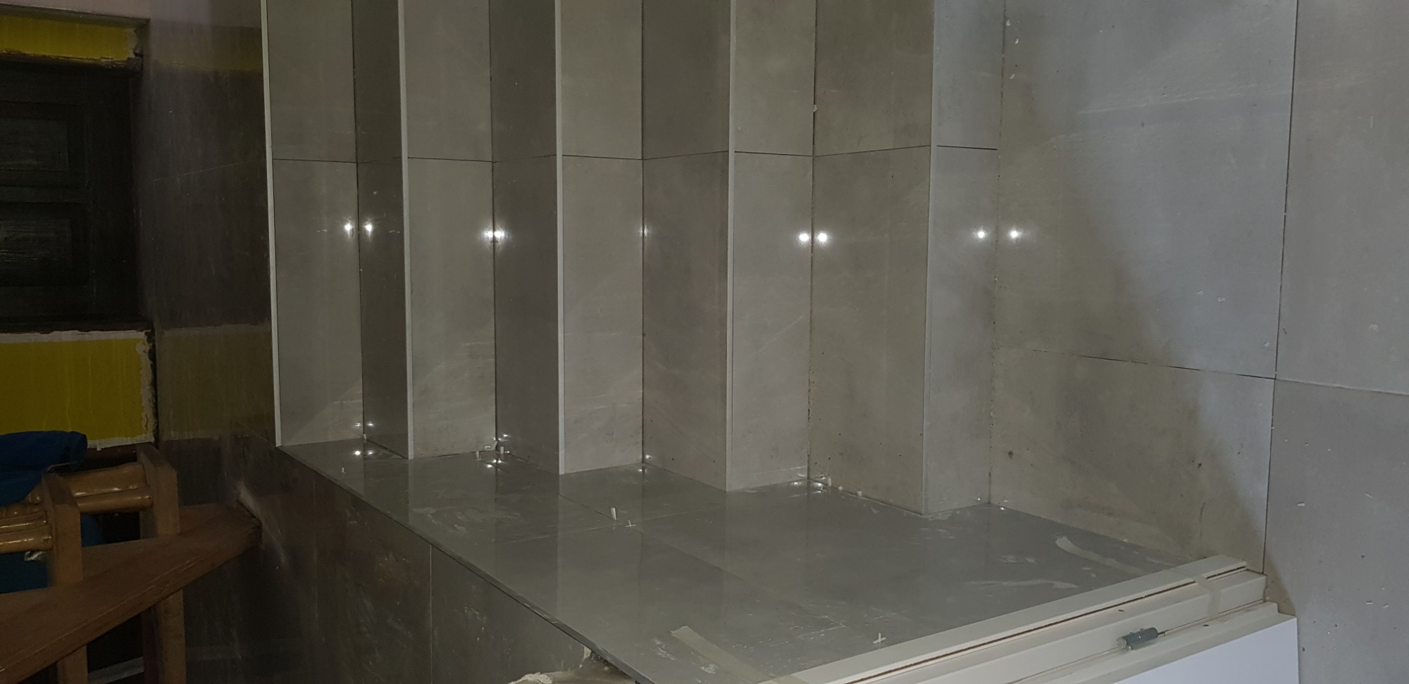 Świeżo wykafelkowana ściana i podłoga z błyszczących, szarych płytek w trakcie prac wykończeniowych; widoczne resztki kleju i fugi.