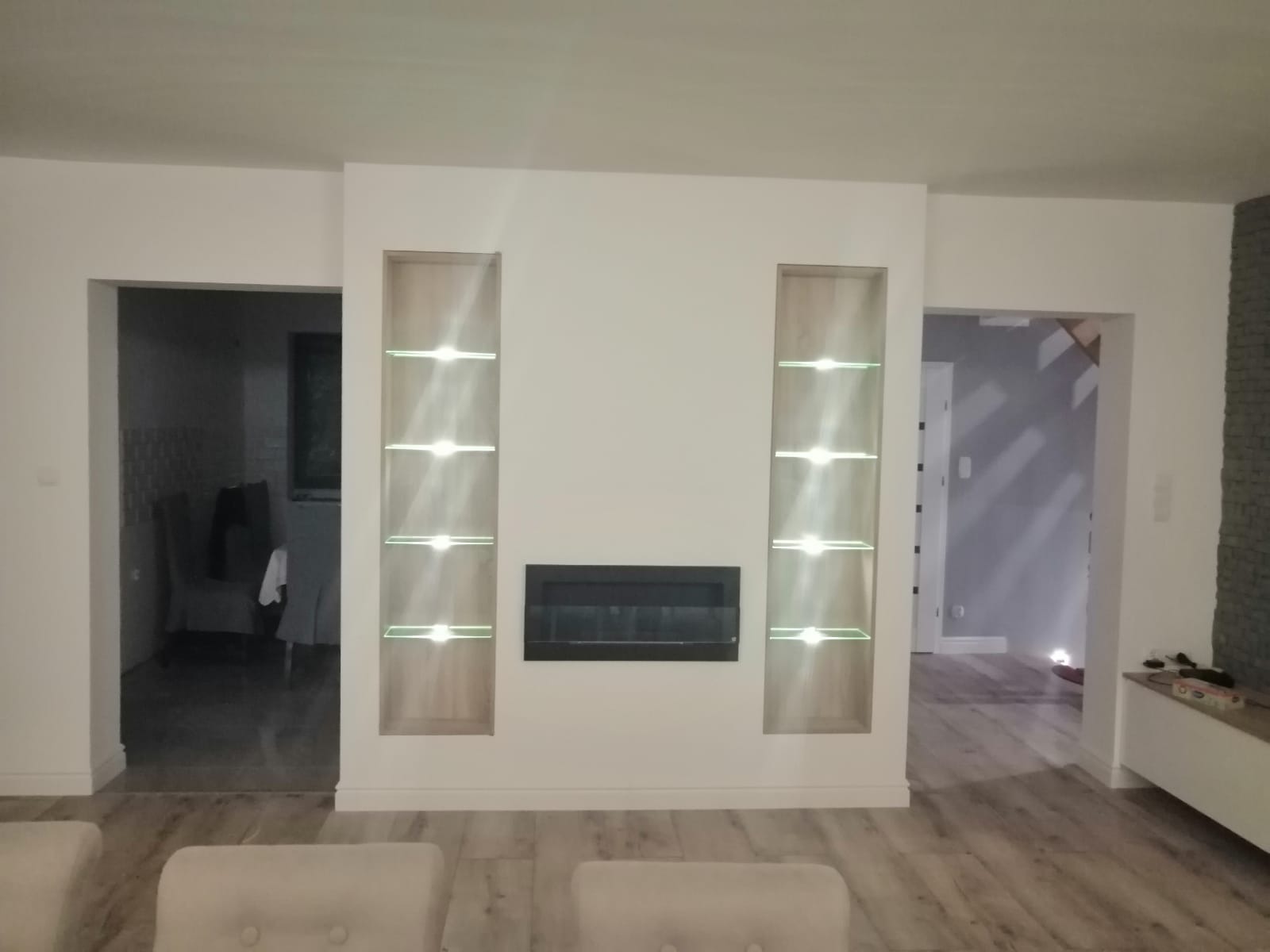 Salon z kominkiem i podświetlanymi półkami w ścianie, widok na jadalnię i korytarz, parkiet imitujący drewno.